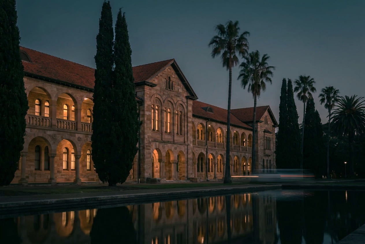 A Mediterranean university campus building at dusk, warm windows lit from inside.